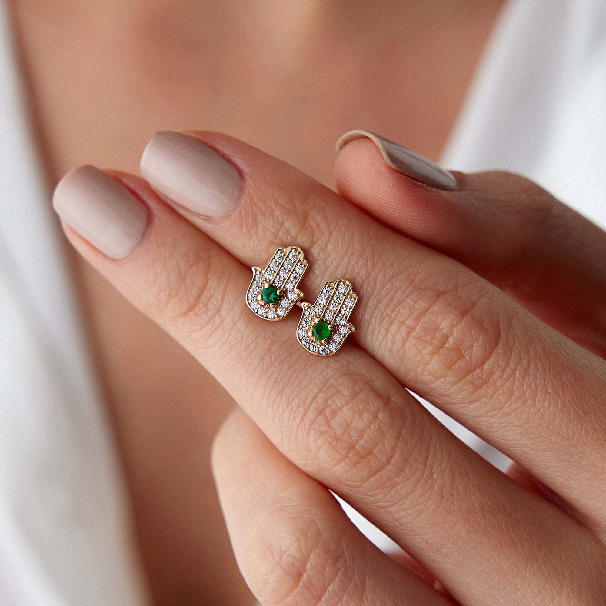 Hand holding a pair of hamsa studs with green central stones