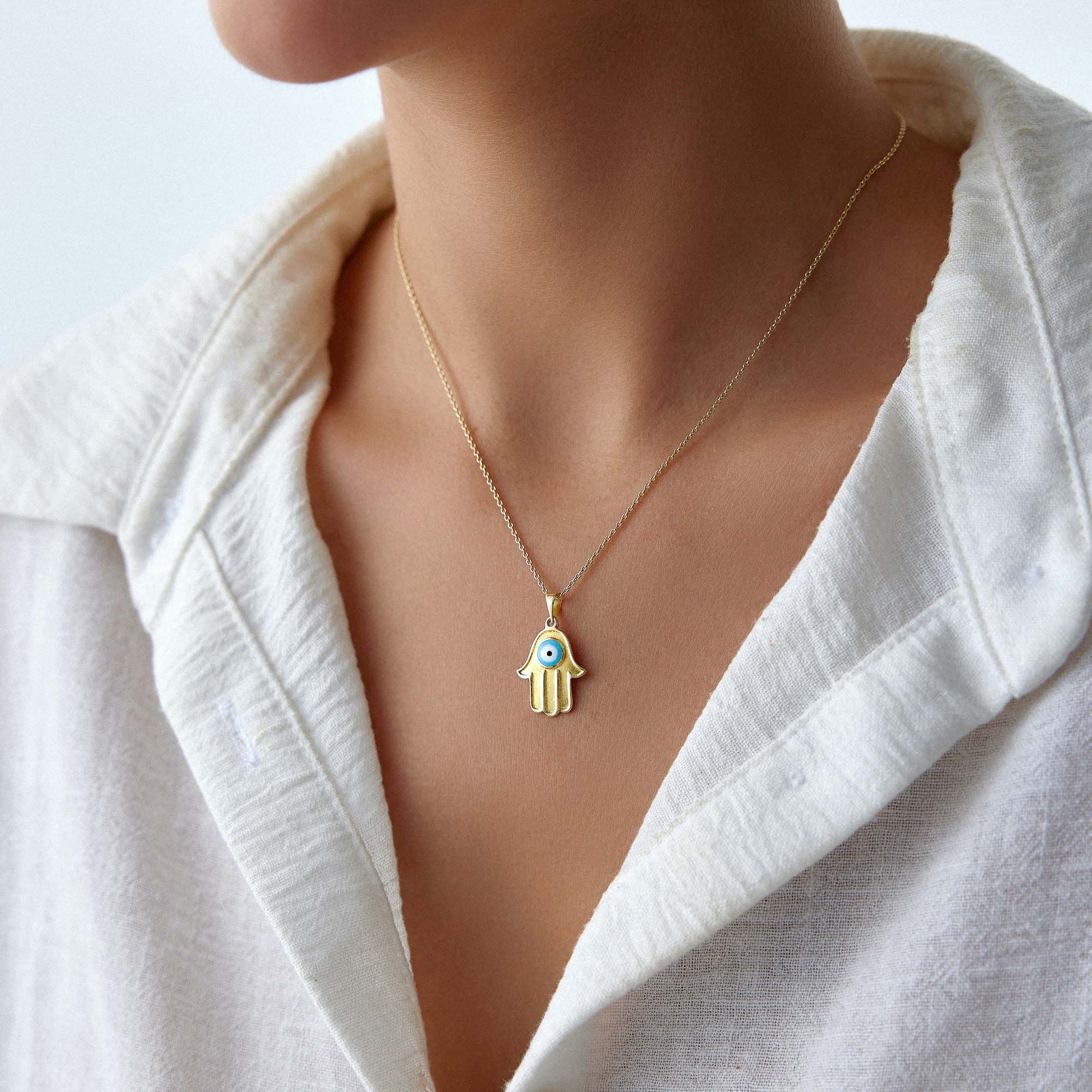 A 14k yellow gold necklace with a hamsa hand pendant featuring a turquoise evil eye, worn by a person.
