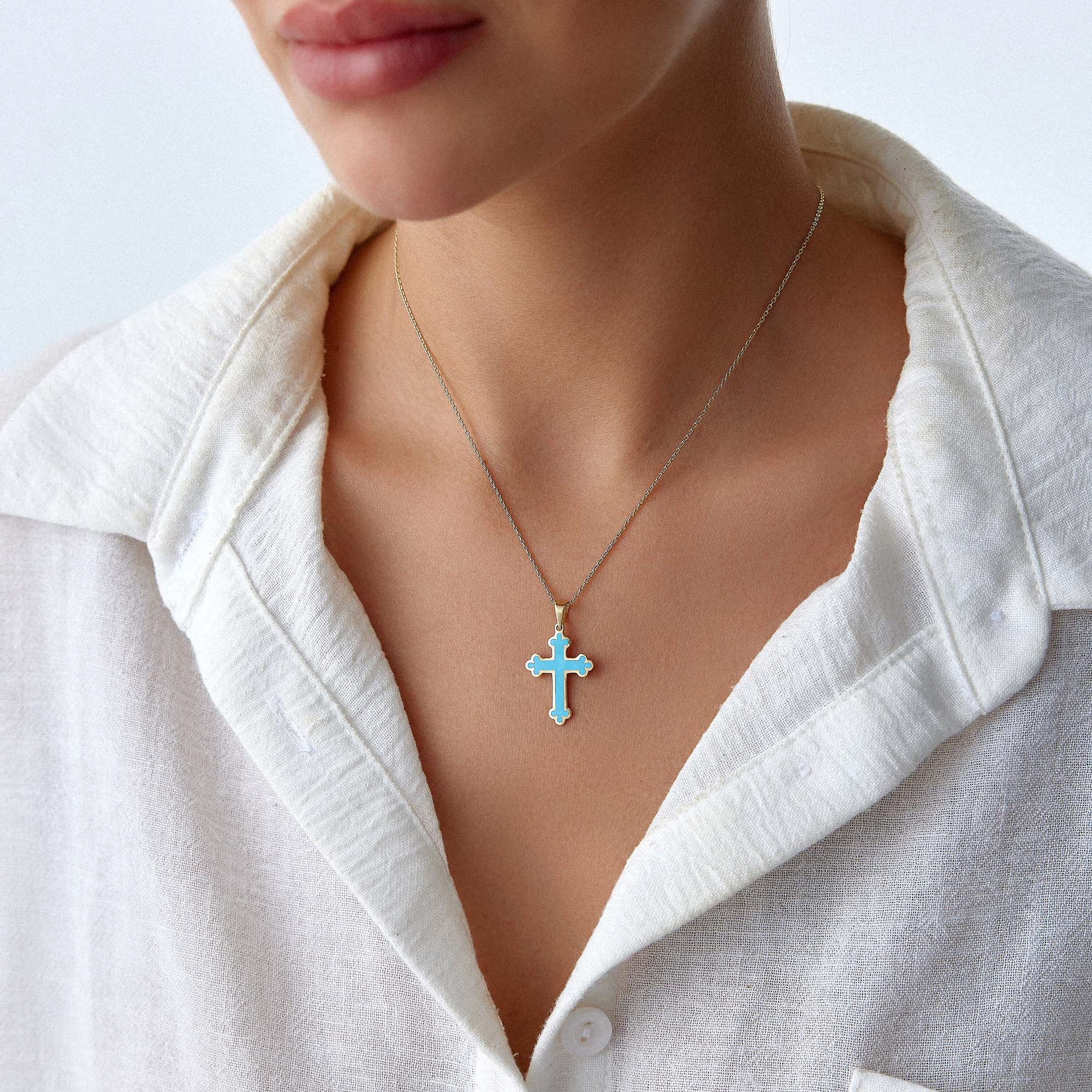Close-up of a 14k yellow gold large ocean blue cross pendant on a gold chain worn by a person in a white shirt.