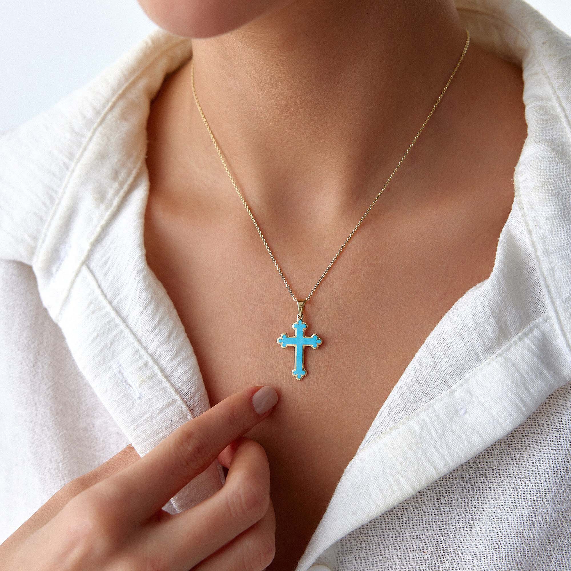 Close-up of a 14k yellow gold large ocean blue cross pendant on a gold chain worn by a person in a white shirt.