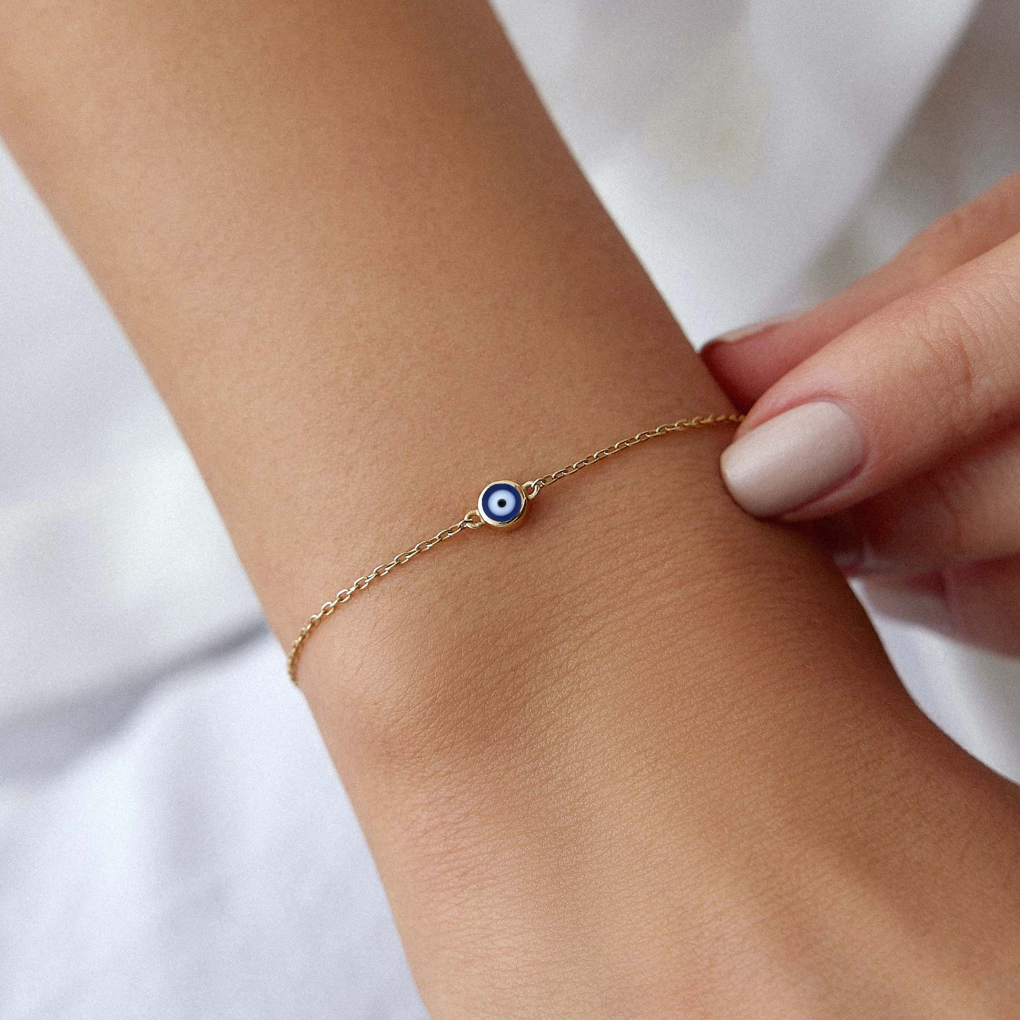 14K Yellow Gold bracelet with a navy blue evil eye charm on a wrist against a blurred background