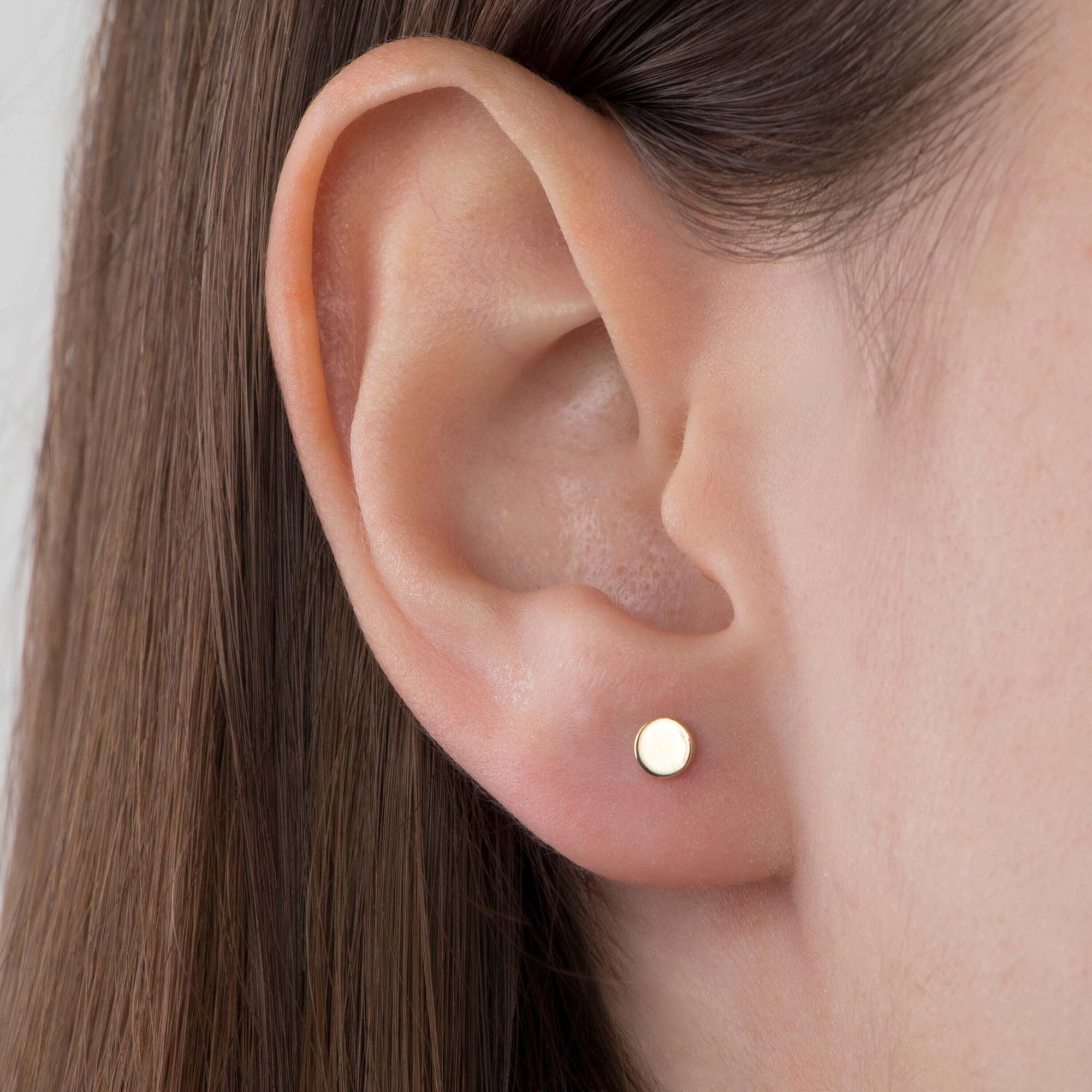 Close-up of an ear wearing a mini circle gold stud earring with a plain background