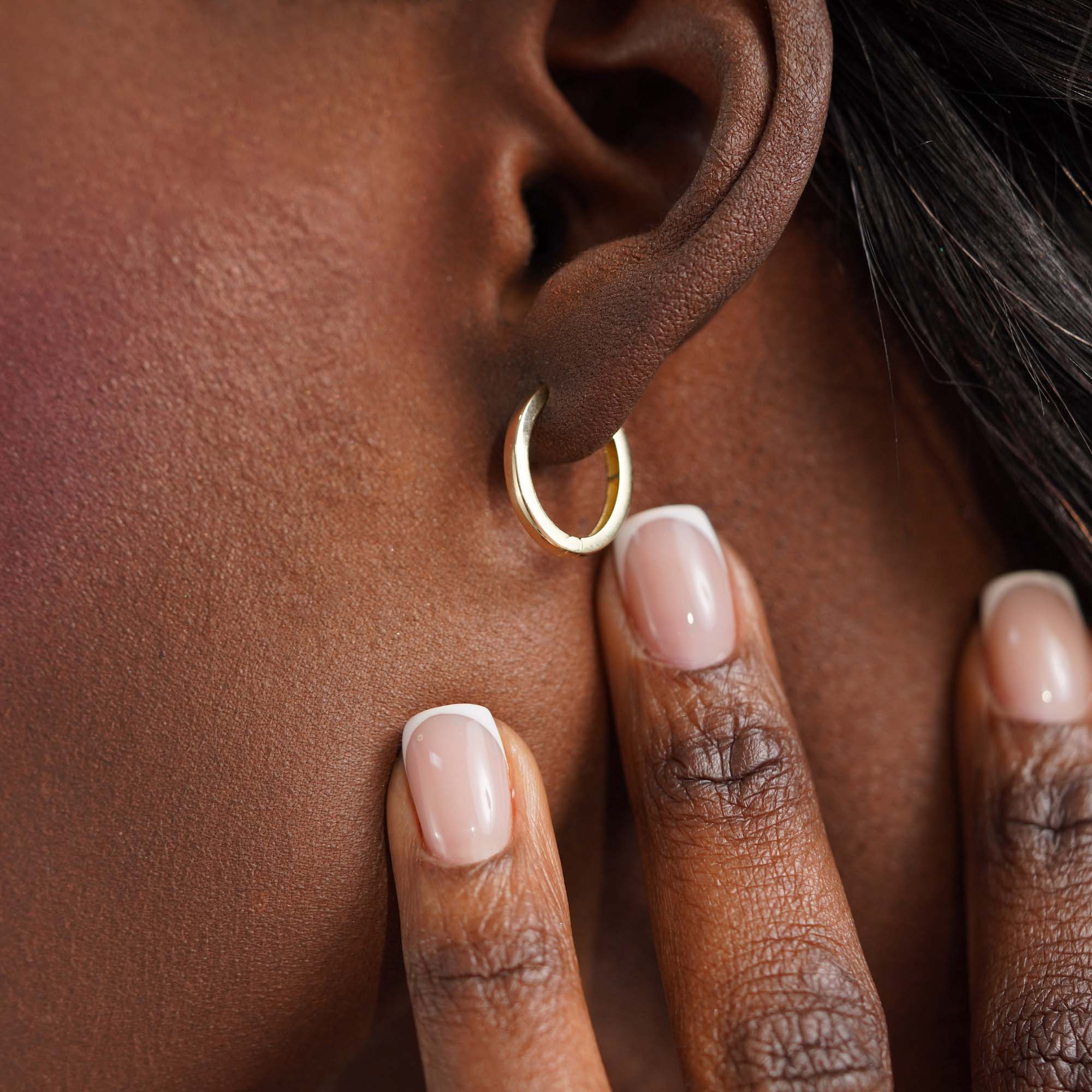 Close-up of an ear wearing an oval shaped gold hoop earring with a hand touching the skin.