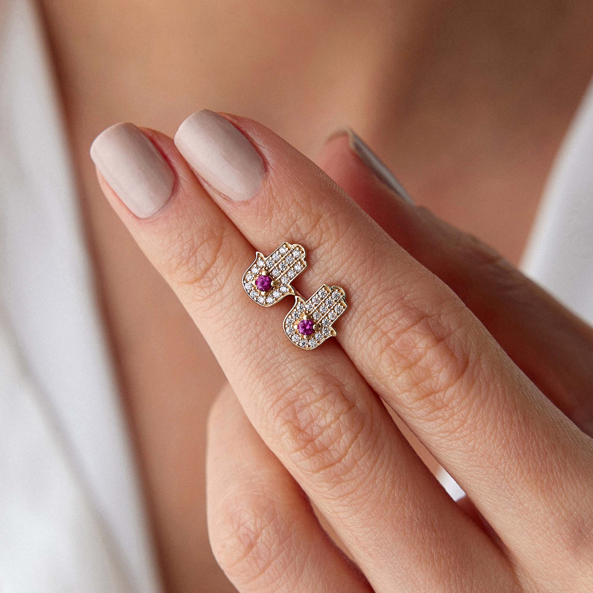 Hamsa hand earrings with pink gemstones on a finger against a neutral background