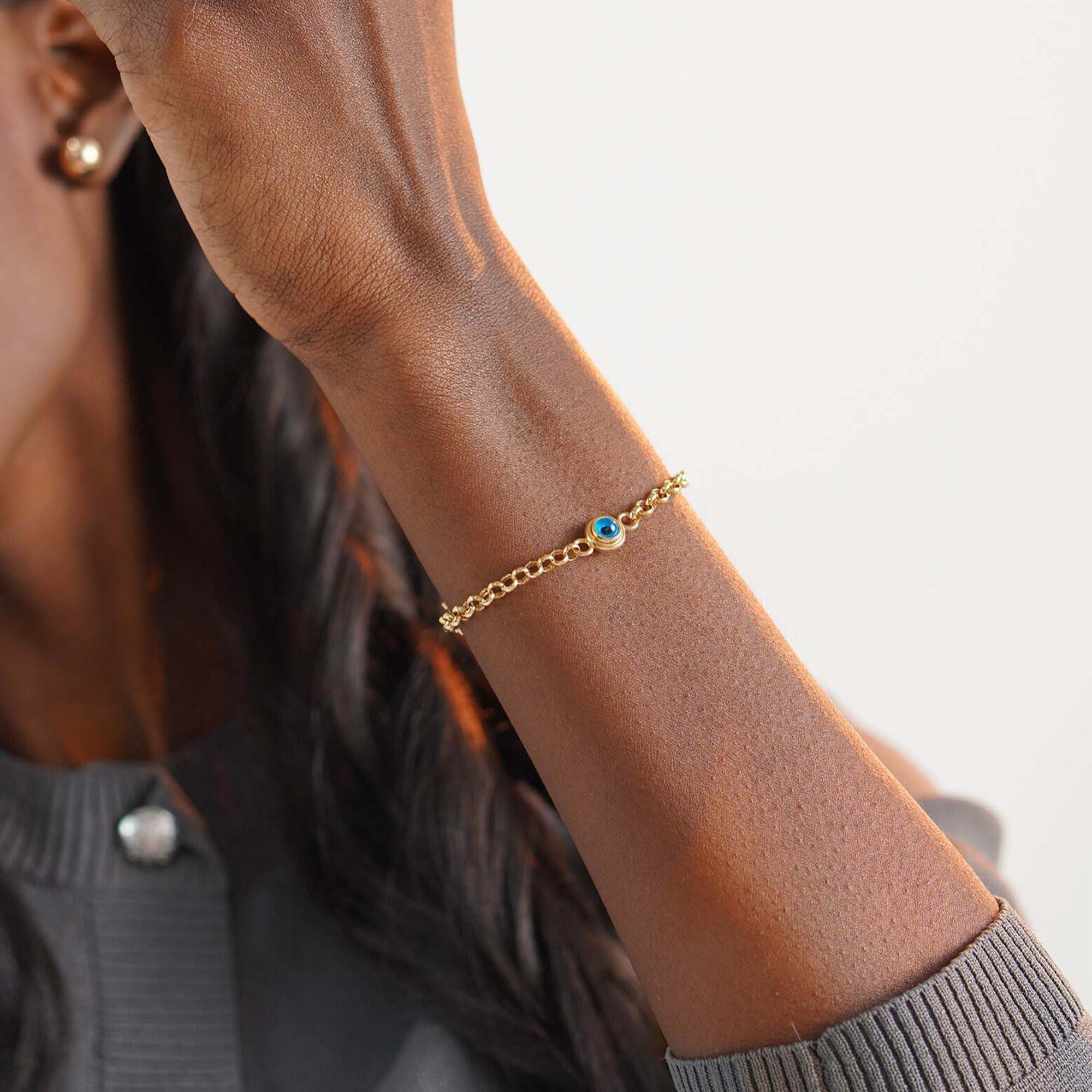 Gold bracelet with a blue evil eye on a person's wrist against a neutral background
