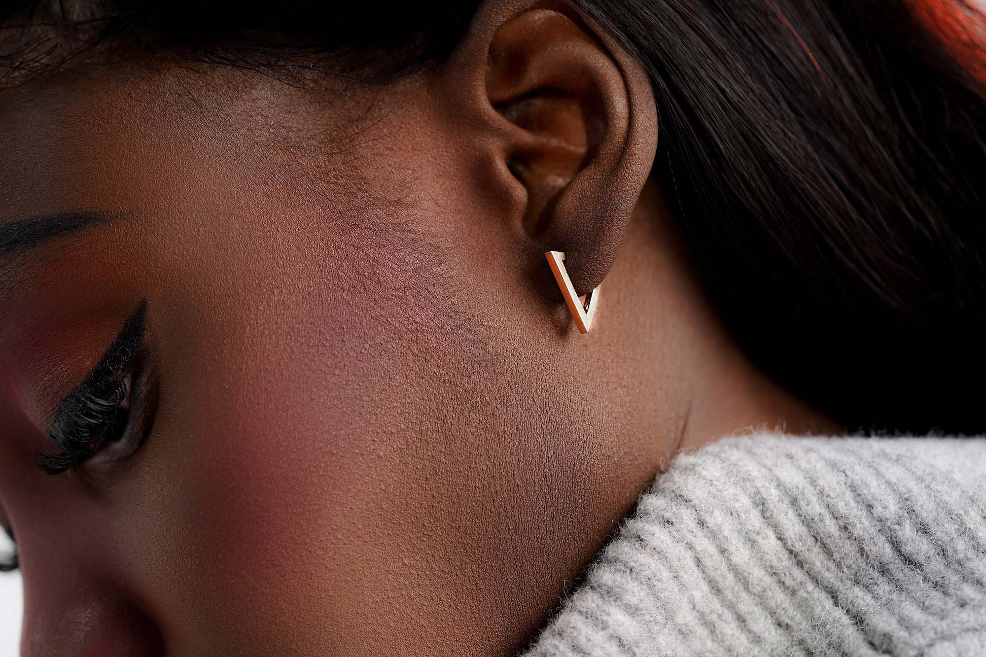 Close-up of a person wearing a triangle shaped gold hoop earring with a neutral background