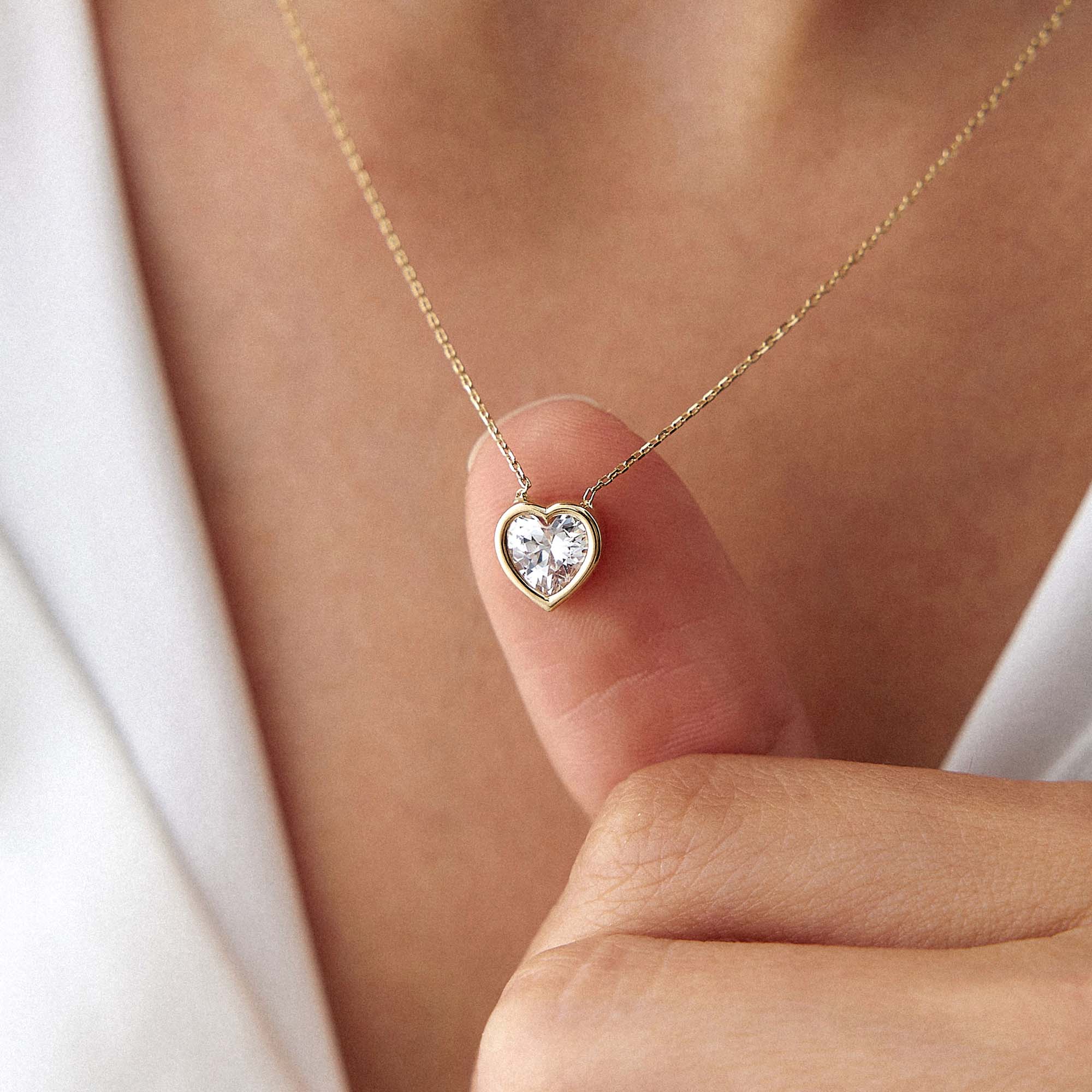 Heart-shaped cubic zirconia pendant on a gold chain held by a hand against a white background