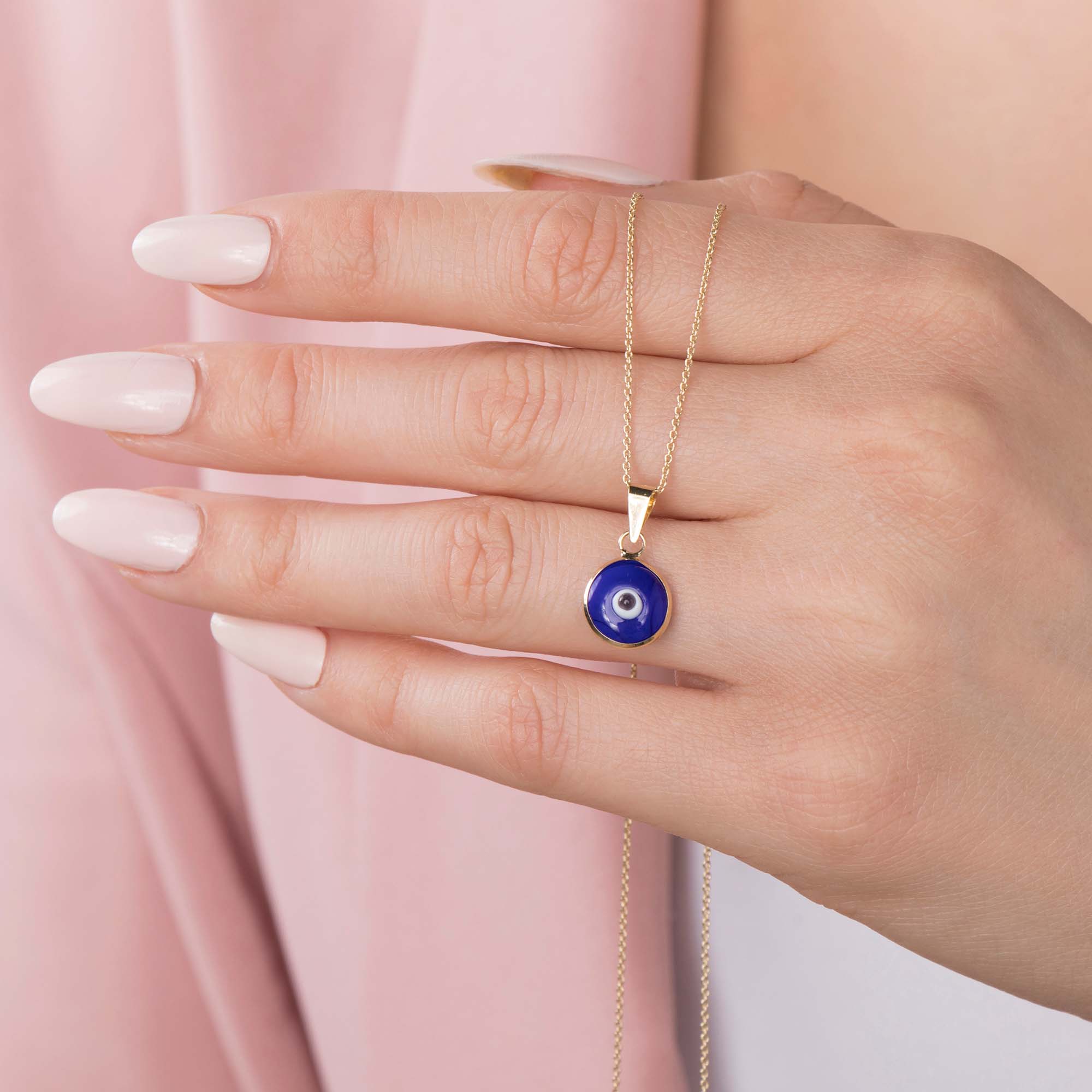 Hand holding a gold chain with a navy blue evil eye pendant against a pink background