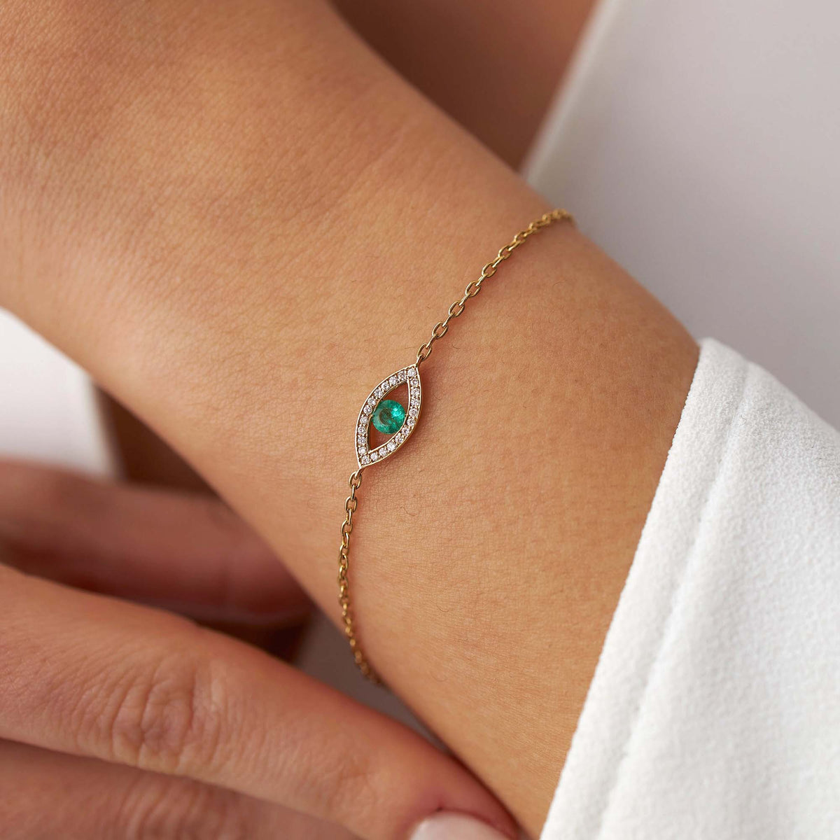 A delicate gold chain bracelet with an eye-shaped centerpiece featuring a green emerald and small clear diamonds, shown worn on a person's wrist.