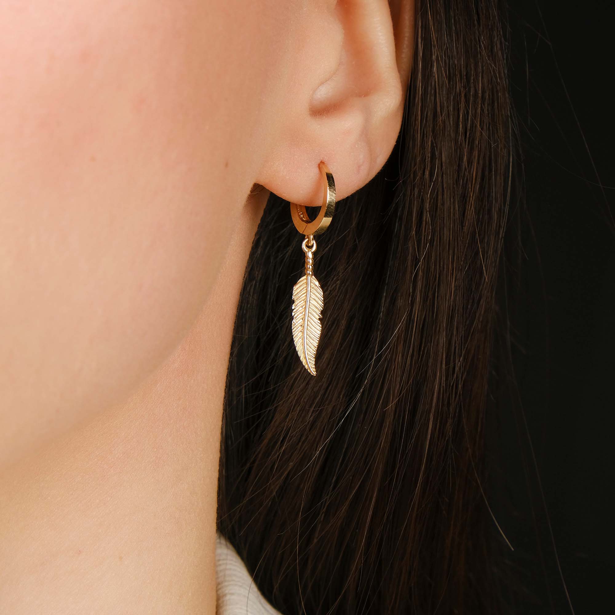 Gold hoop earring with a feather charm worn by a person.
