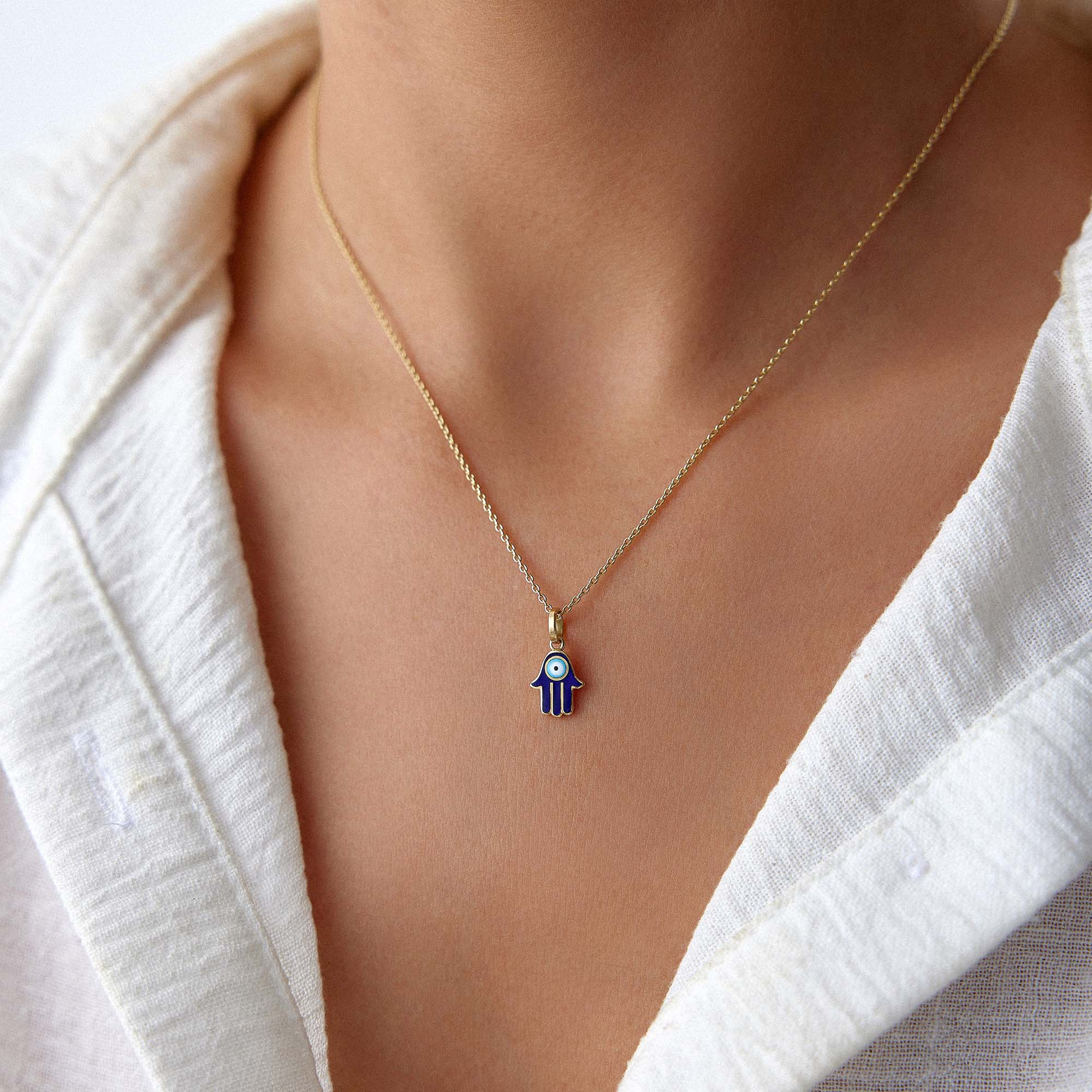 A blue hamsa pendant with an evil eye center worn on a thin gold chain by a model on a white shirt.