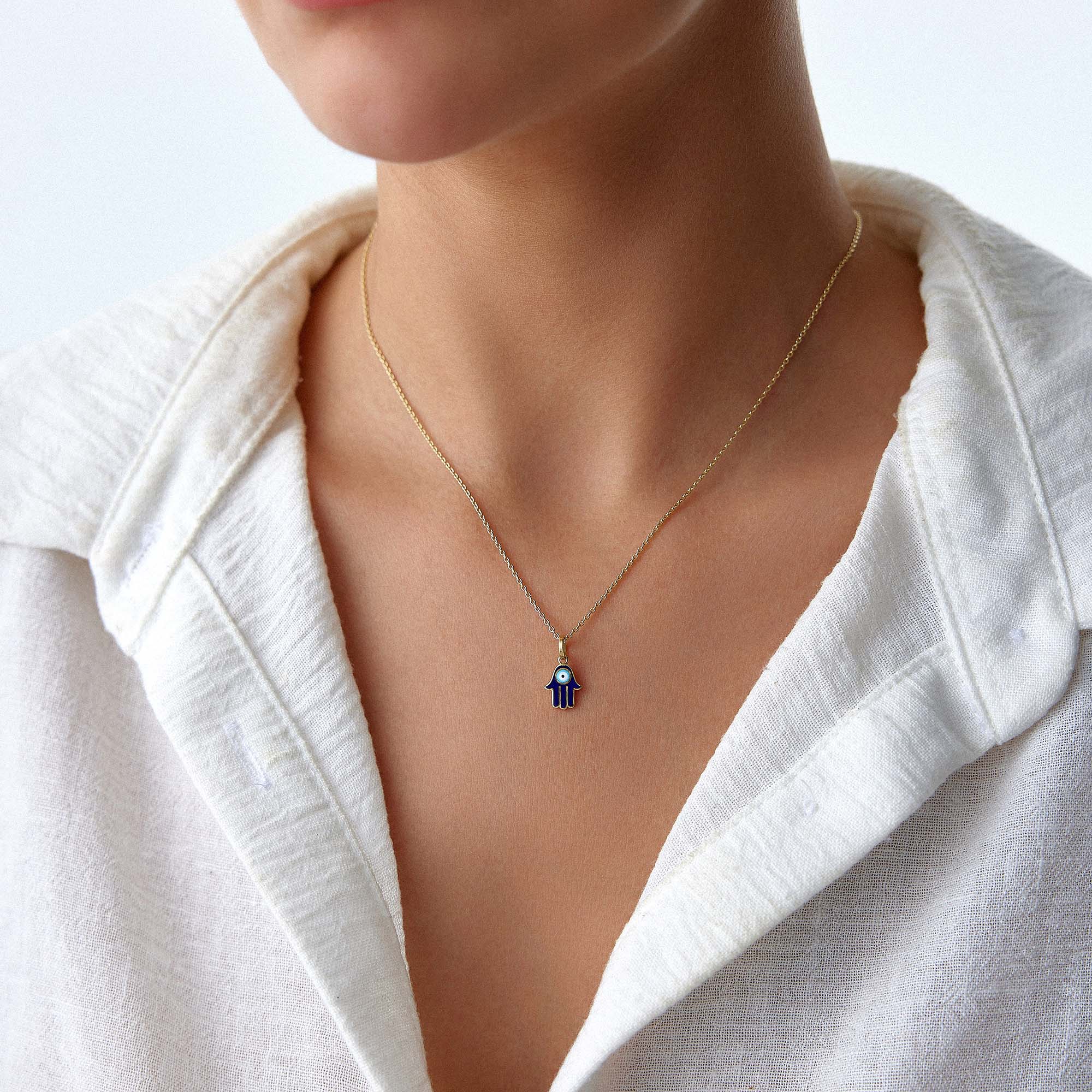 A blue hamsa pendant with an evil eye center worn on a thin gold chain by a model on a white shirt.