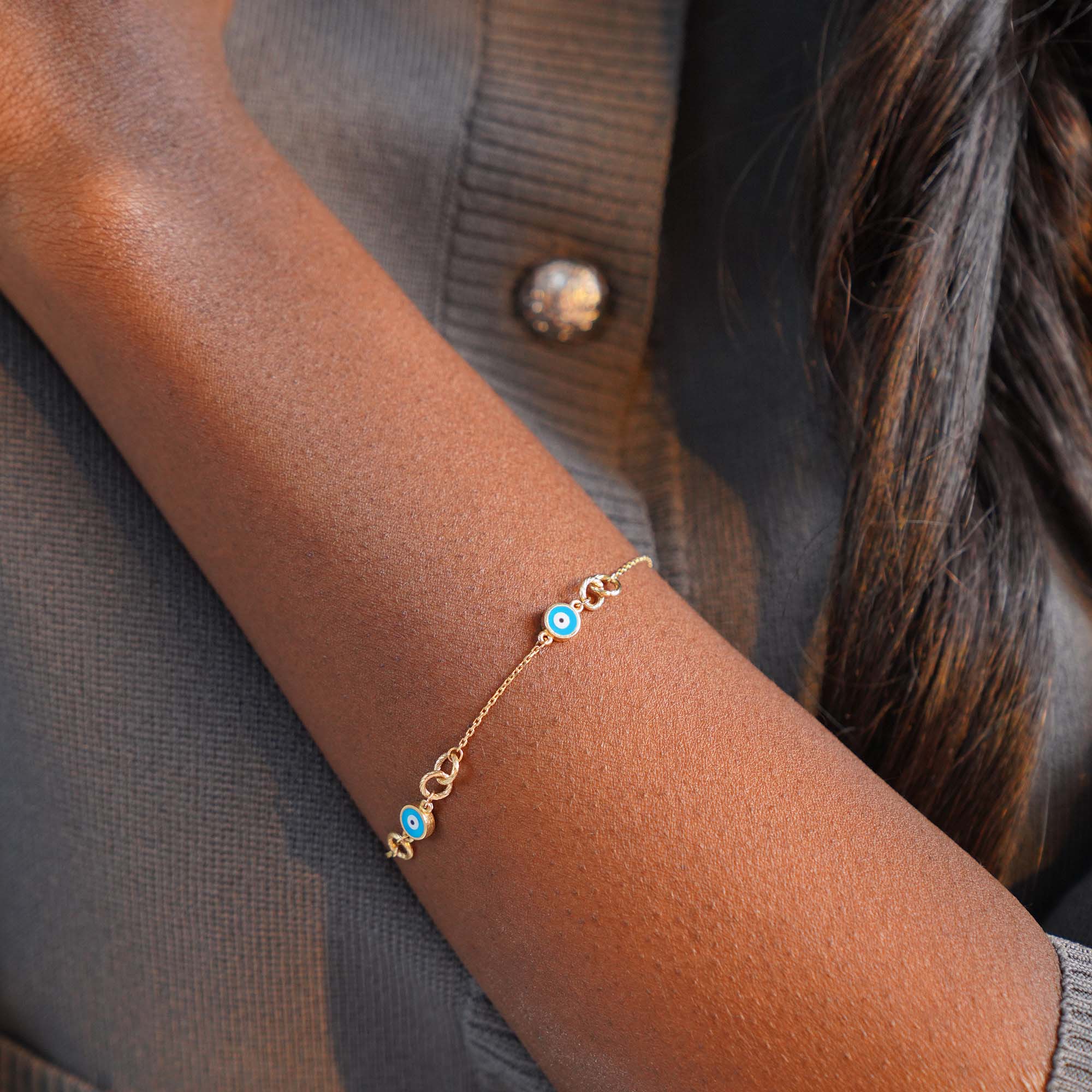 Gold bracelet with a blue evil eyes on a person's wrist against a light gray background