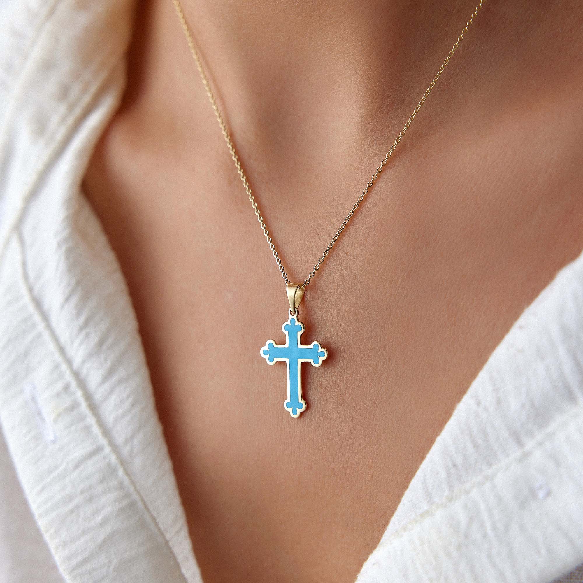 Close-up of a 14k yellow gold large ocean blue cross pendant on a gold chain worn by a person in a white shirt.