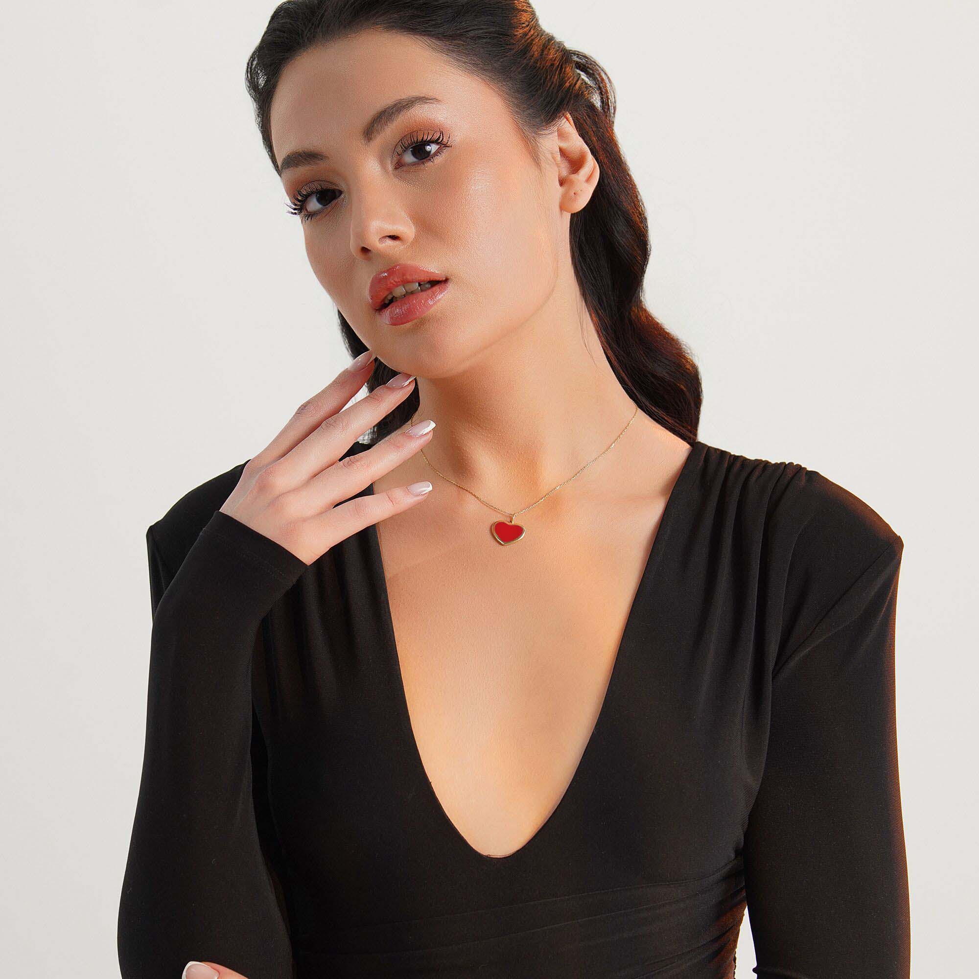 Woman wearing a black outfit with a deep neckline and a large red heart-shaped pendant necklace on a light gray background