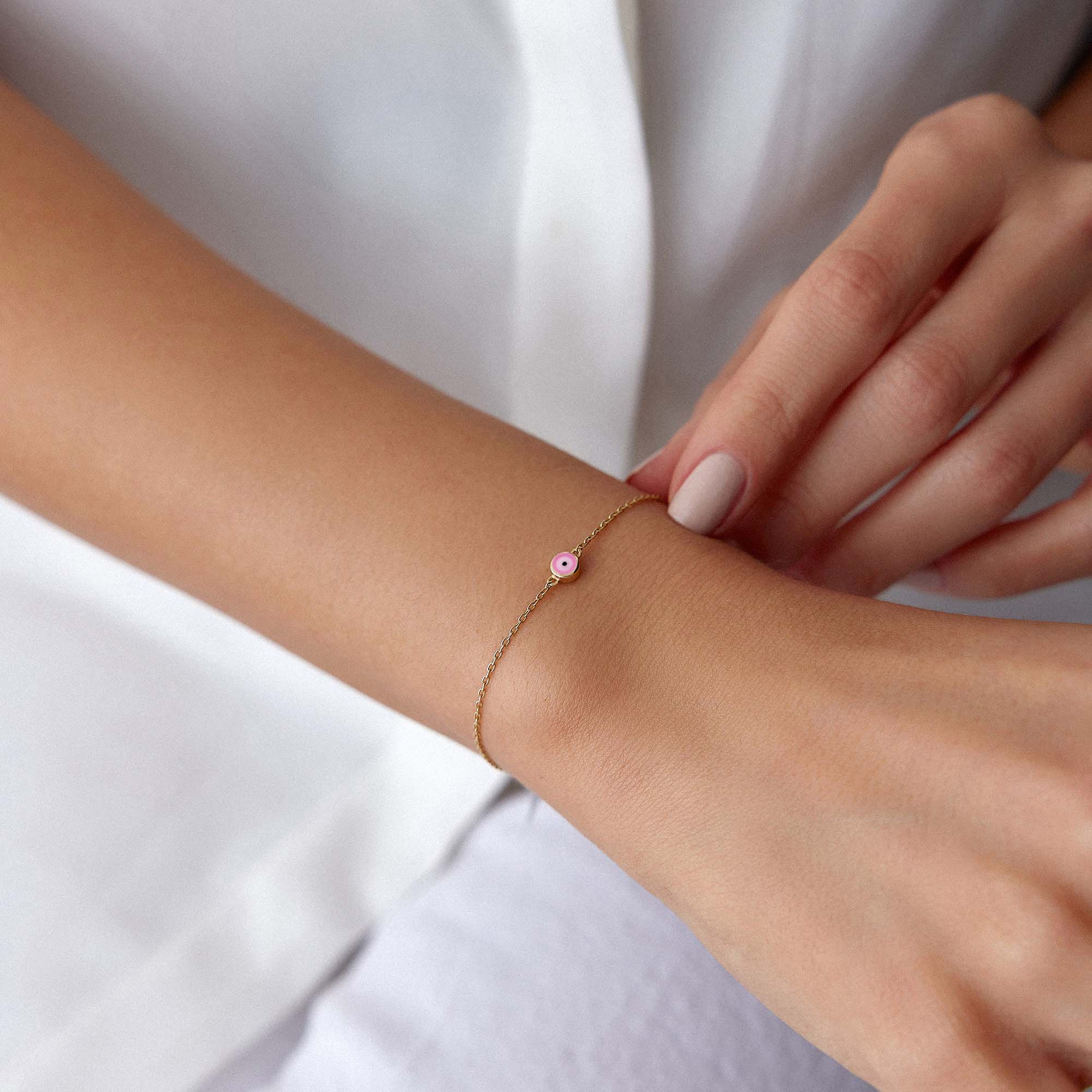Close-up of a wrist wearing a delicate bracelet with a small pink evil eye charm against a white background.