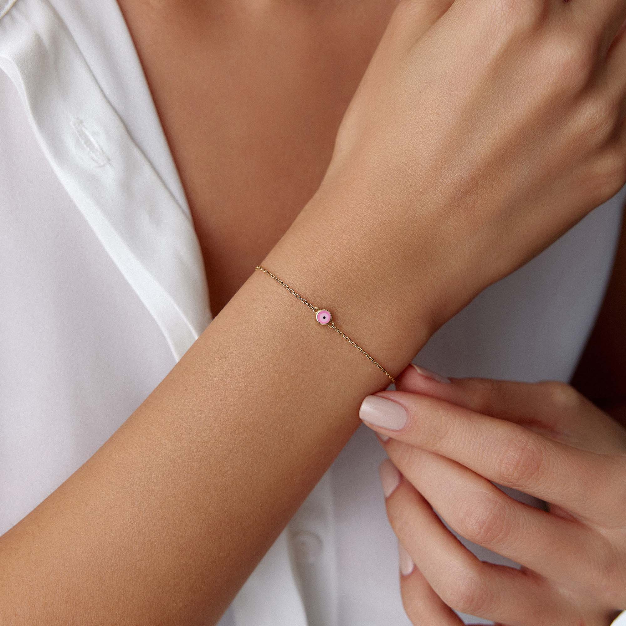 Close-up of a wrist wearing a delicate bracelet with a small pink evil eye charm against a white background.