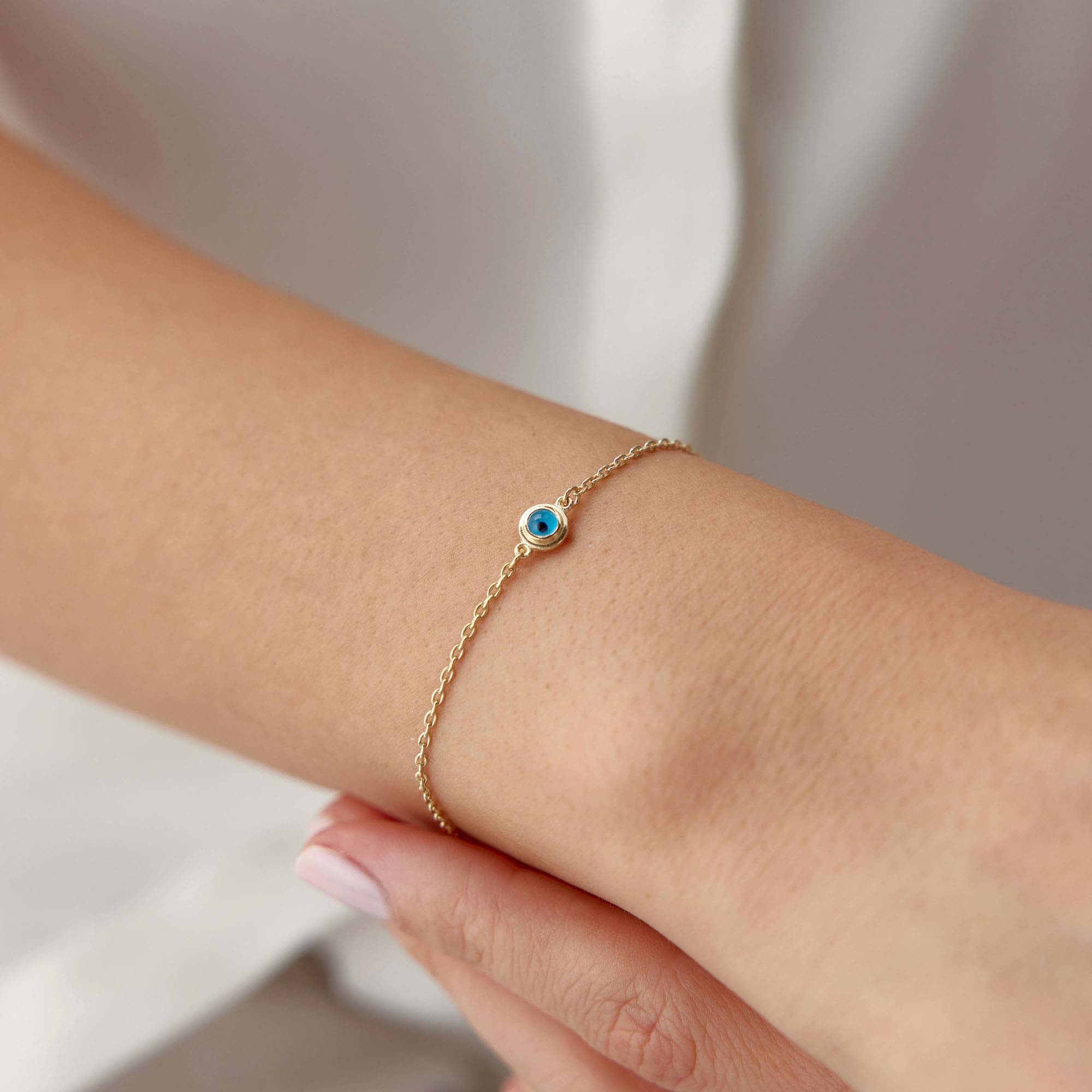 Gold bracelet with a minimal blue evil eye charm on a wrist against a neutral background