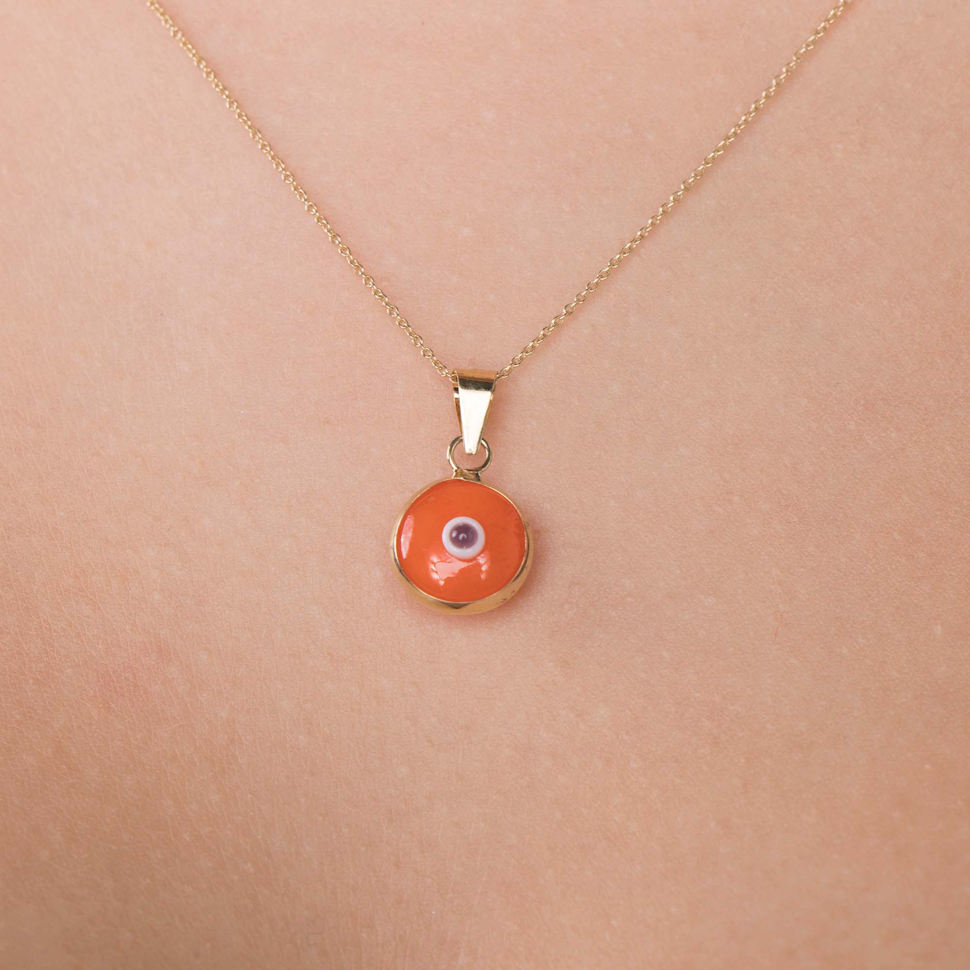 A model wearing an orange evil eye pendant on a thin gold chain shown up-close
