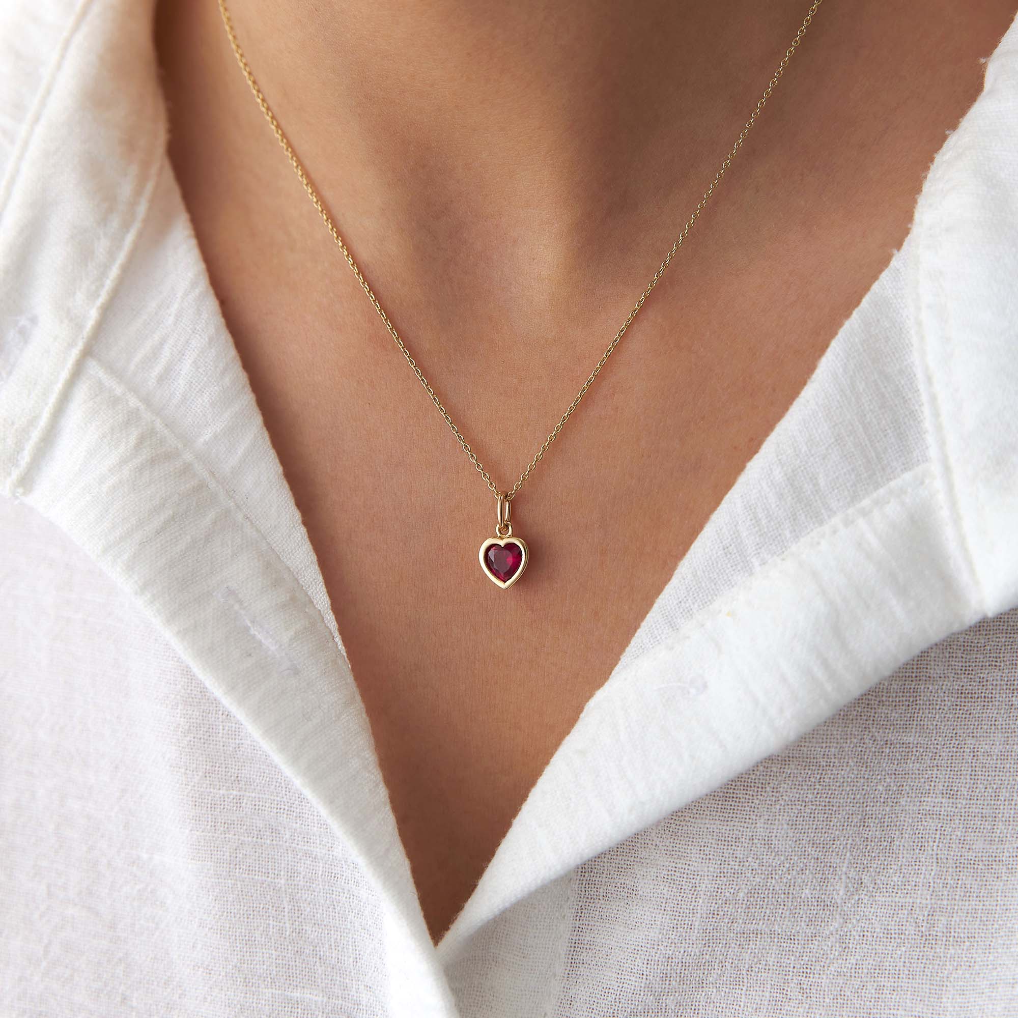 Gold necklace with a red heart-shaped pendant worn by a person in a white shirt.