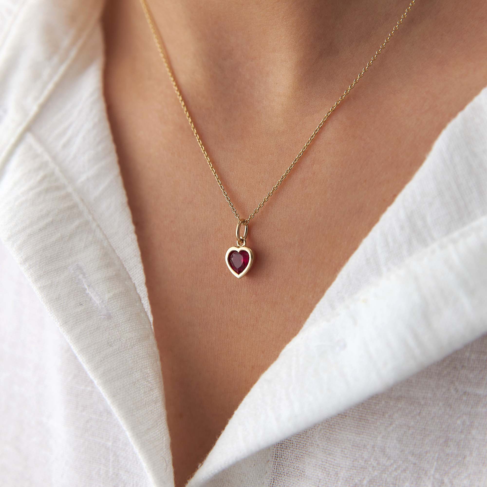 Gold necklace with a red heart-shaped pendant worn by a person in a white shirt.