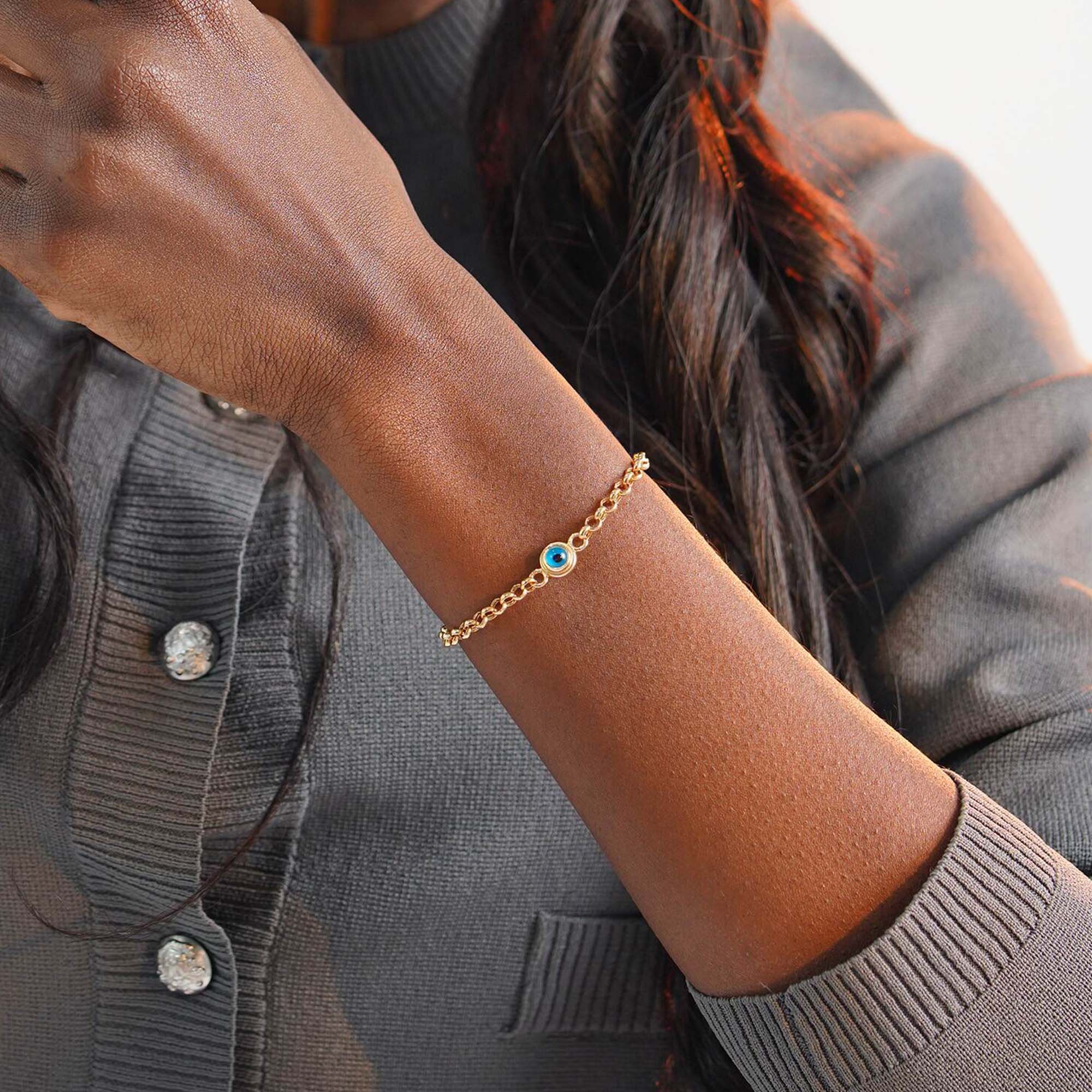 Close-up of a person wearing a gold bracelet with a blue evil eye on a gray cardigan background.