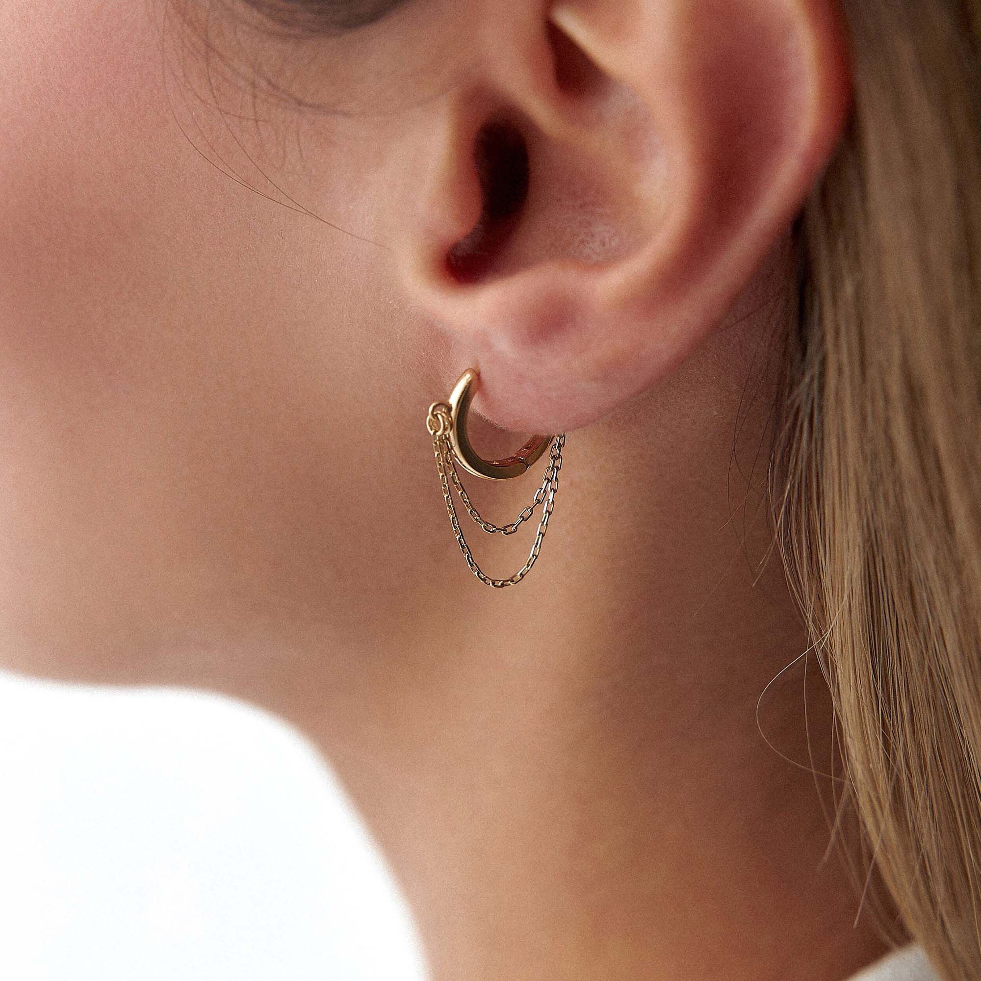 A close-up image of a woman's ear wearing a 14k yellow gold hoop earring with dangling chain details.