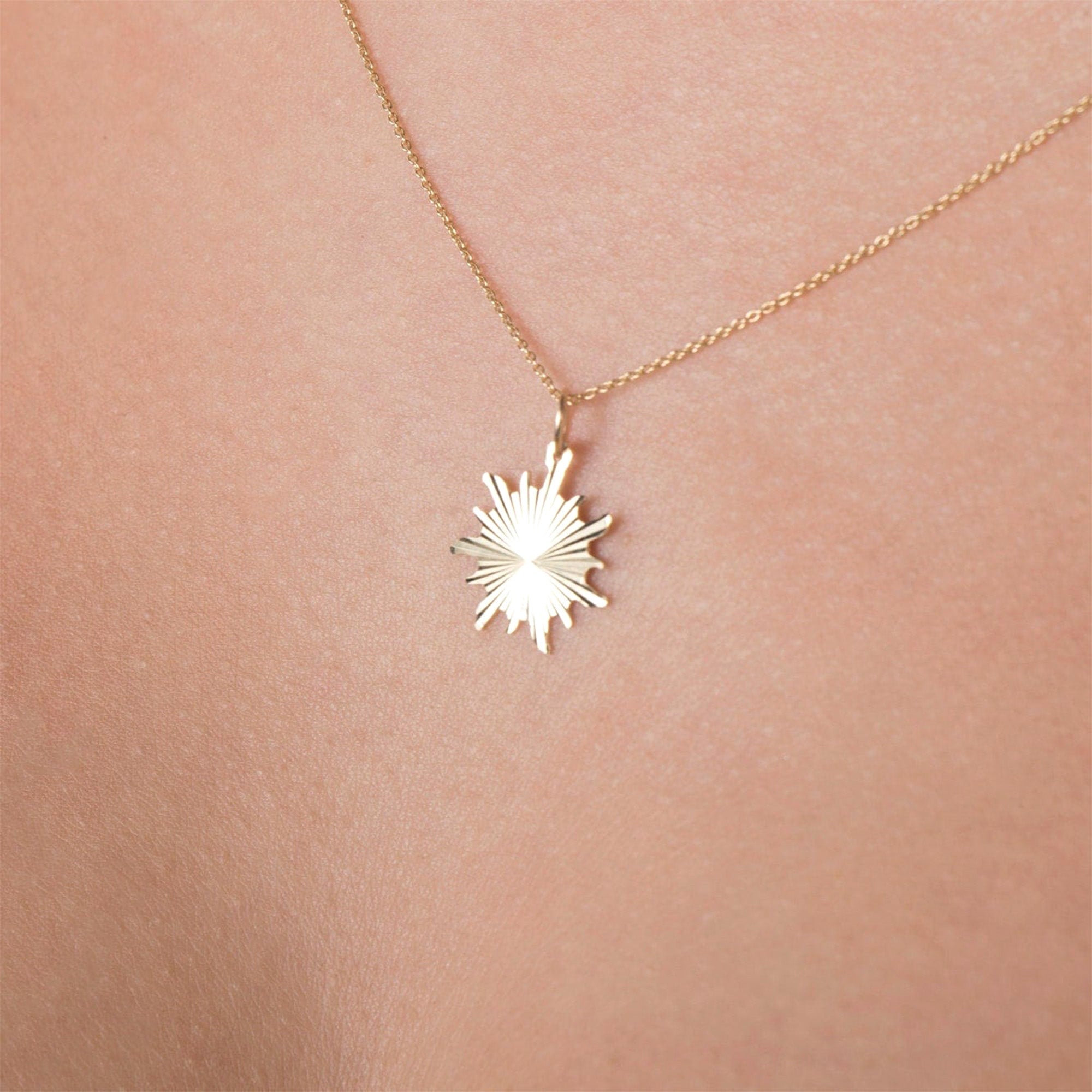 A minimal sunburst necklace in 14K yellow gold, with the pendant displayed against a skin-like background.