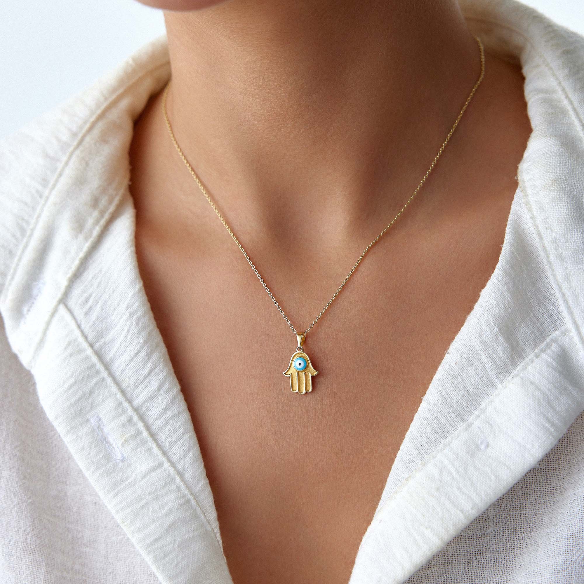 A person wearing a white v-neck shirt showcasing a vintage Hamsa necklace in 14K gold with a turquoise blue evil eye center.